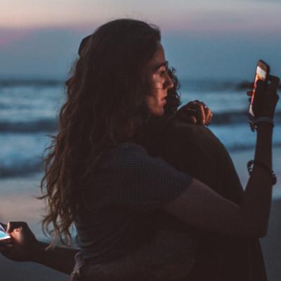 Pareja revisando cada una por su cuenta el móvil, mientras se abrazan en la playa